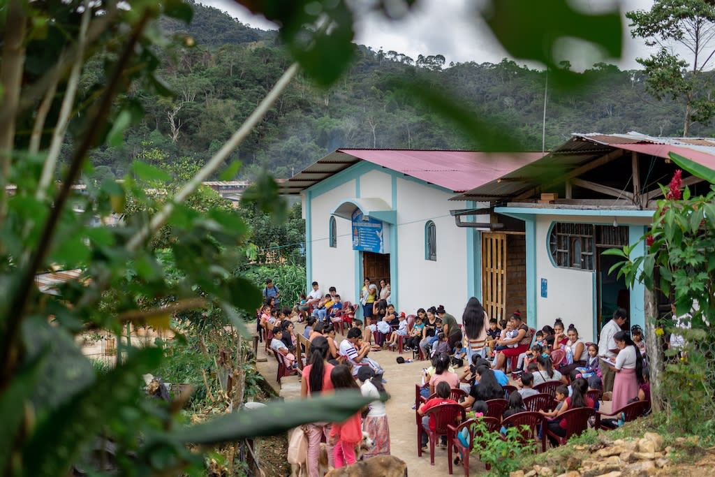 A faraway photo of a Compassion centre. It is surrounded by trees.There are many moms and babies and kids sitting on the outside of the centre.