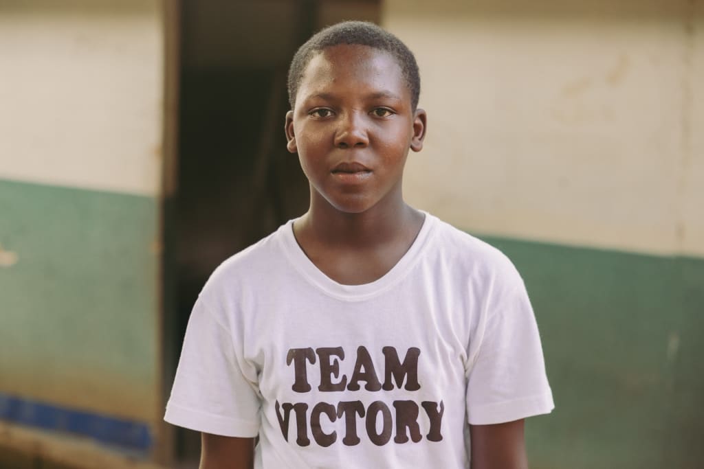 A young boy stands looking into the camera wearing a white t-shirt that reads "Team Victory."