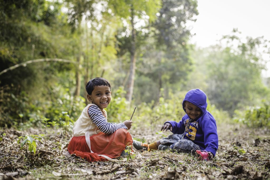 Two young children sit on the ground and hold sticks in the forest.