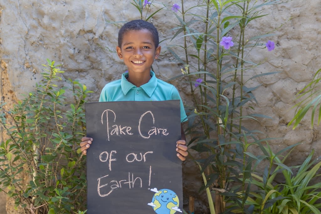A boy in a turquoise shirt stands in the grass against a stone wall holding a sign that says, "Take care of our earth!"