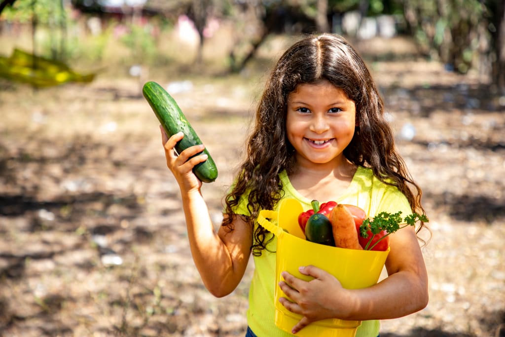 A girl in a yellow tshirt holds a yellow bucket full of vegetables.
