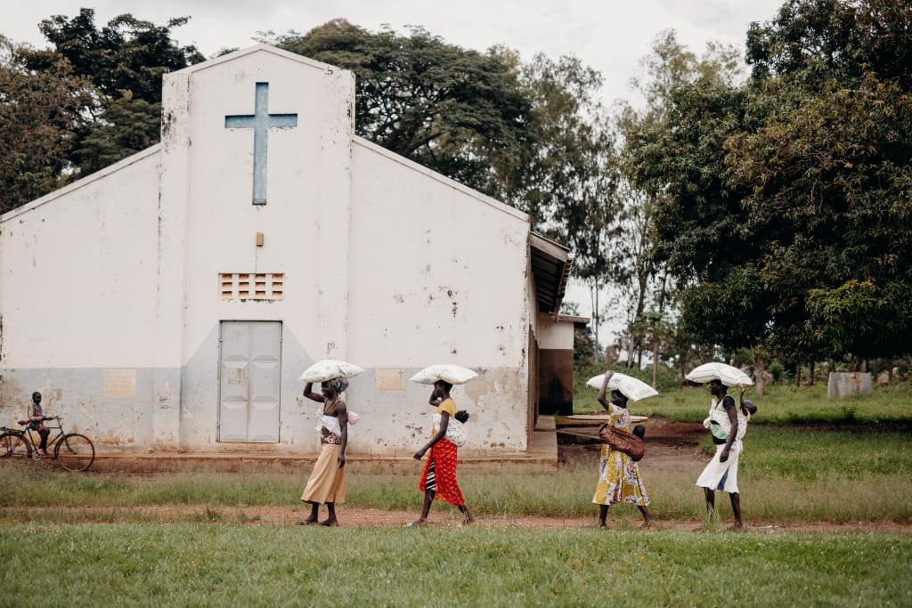 Four women carry bags of grain on their heads, walking in front of a church building.