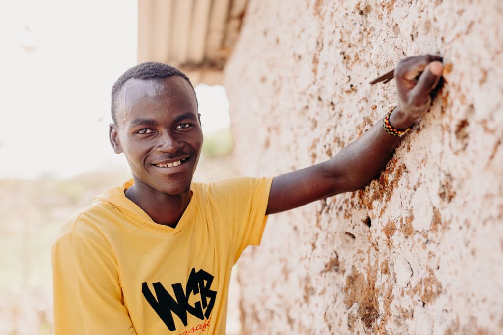 A young man in a yellow t-shirt smiles and rests his arm against a mud wall.