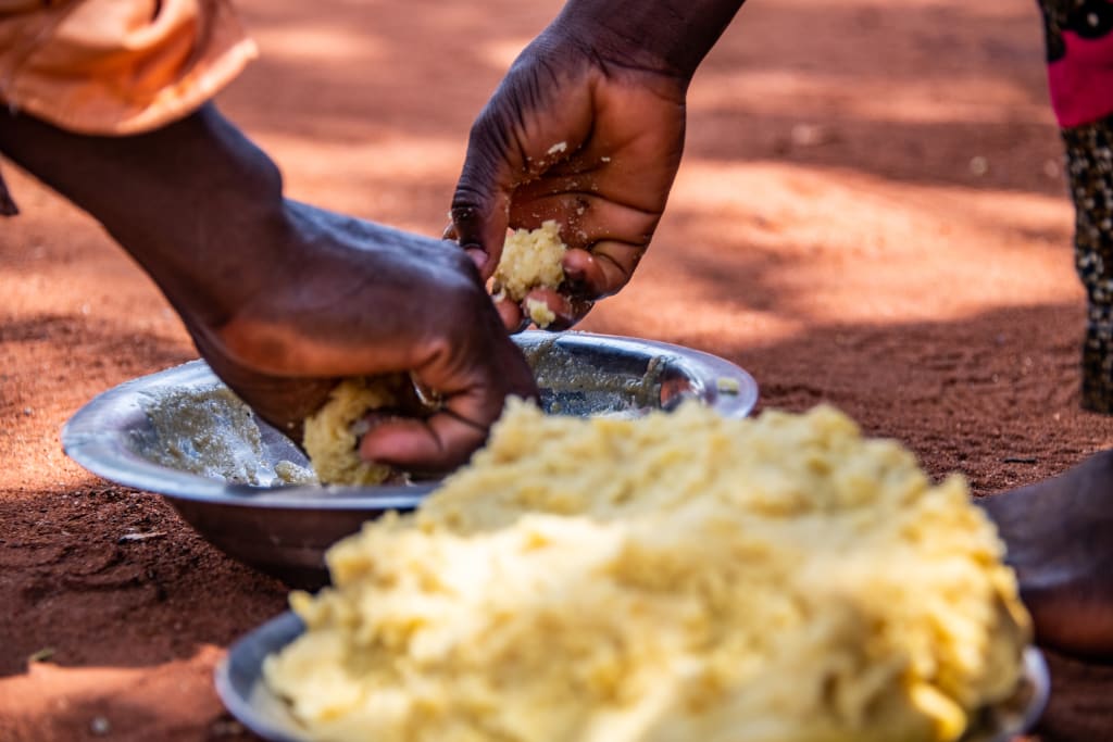 Adult hands working with flour in a bowl on a red dirt ground in Togo.