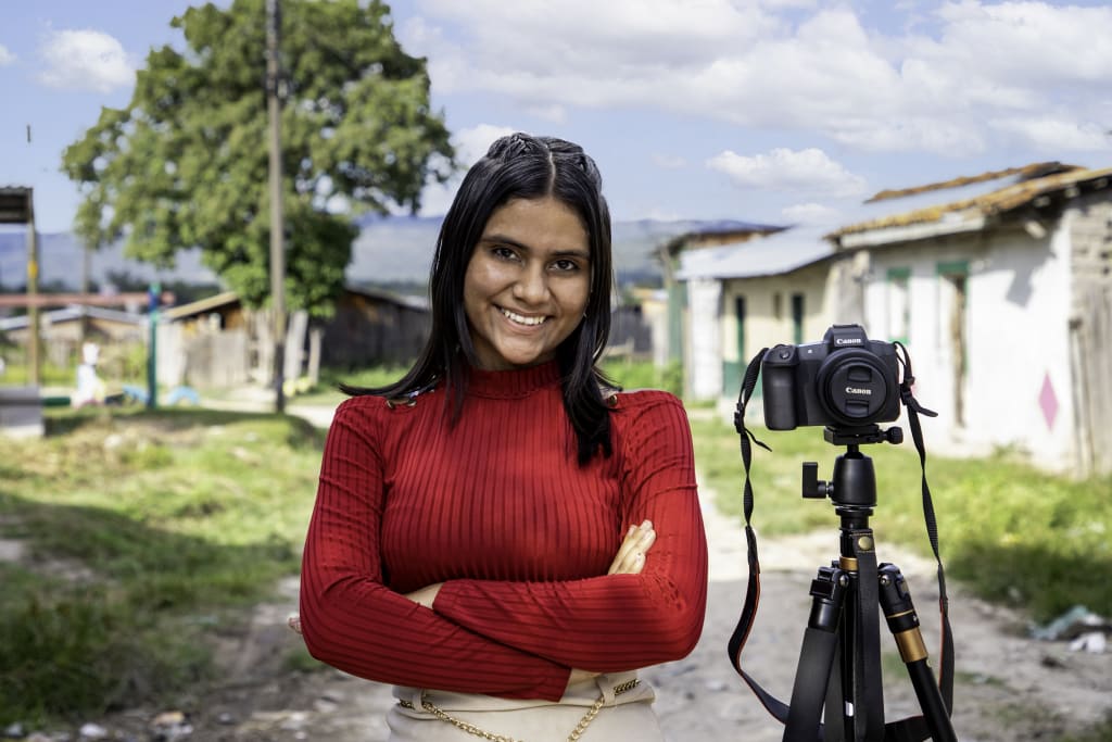 A young woman in a red shirt stands smiling beside a camera on a tripod.
