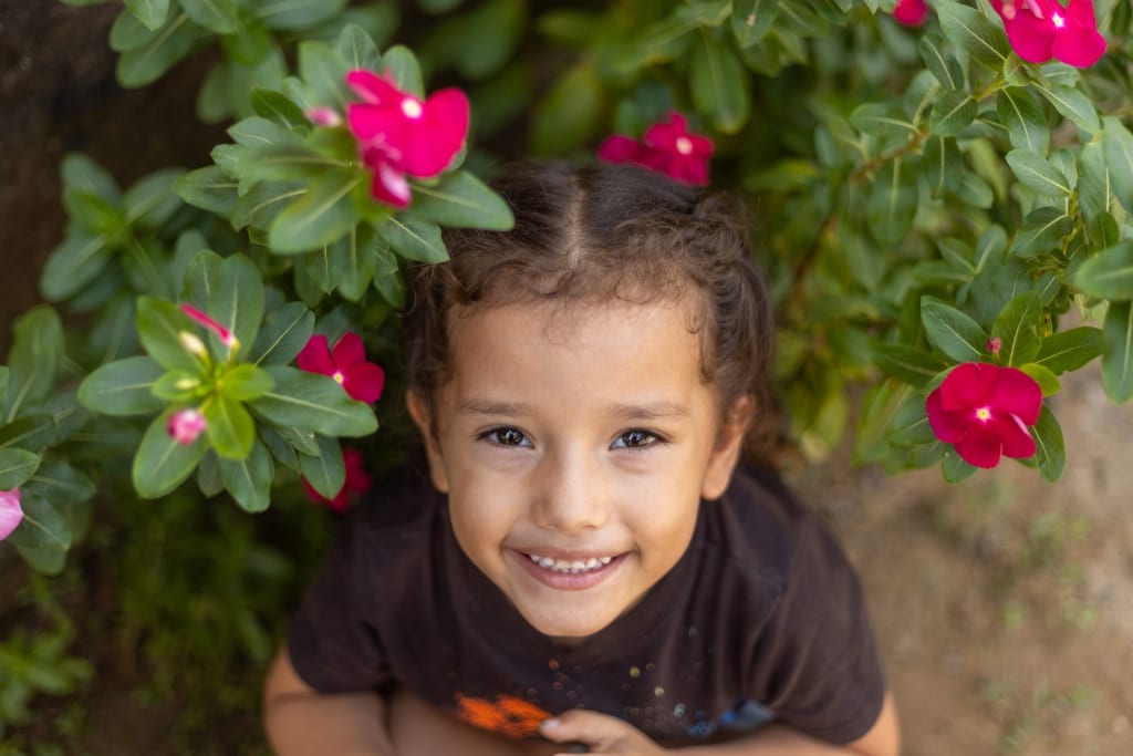A child looking up with a smile. She is standing in front of a bush with blooming flowers.