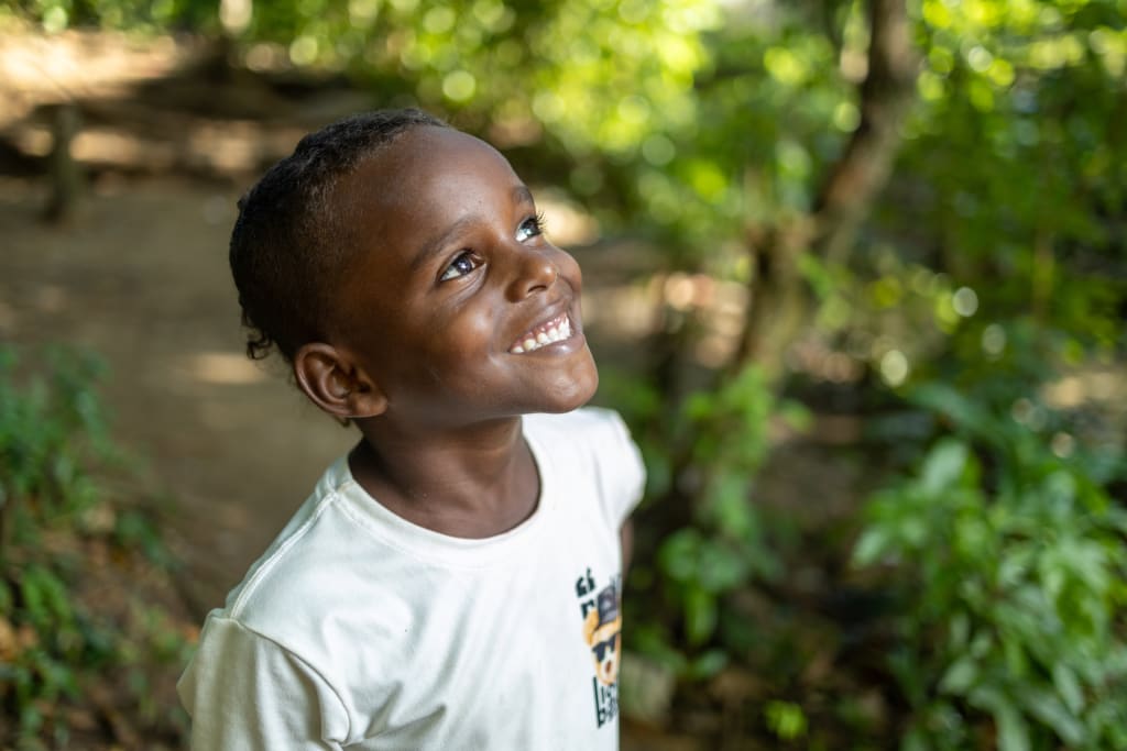 A child looks up with a big smile. He is wearing a teal shirt and standing in the shade of trees.