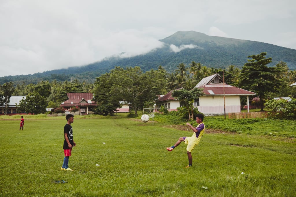Adolescent teen boys wearing black, red, yellow, purple kick a soccer ball in a grass, grassy field. Behind them are homes and a mountain, clouds.
