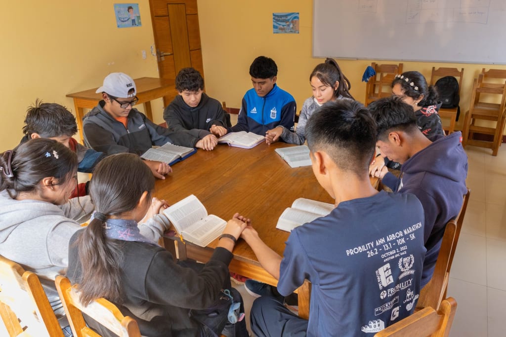Youth sit in a circle at a table with their Bibles, holding hands in prayer.