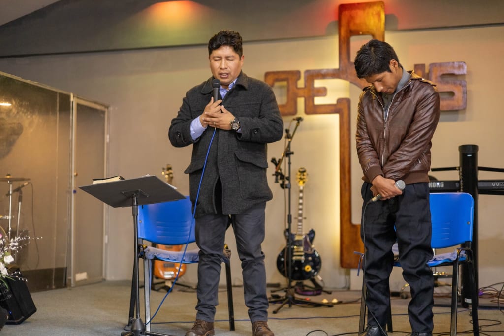 A church leader prays for a preacher. They stand together on stage, the church leader in a black jacket, and the preacher in a brown leather jacket, looking down with their eyes closed. Blue chairs are behind them, along with a Jesus sign and musical instruments.