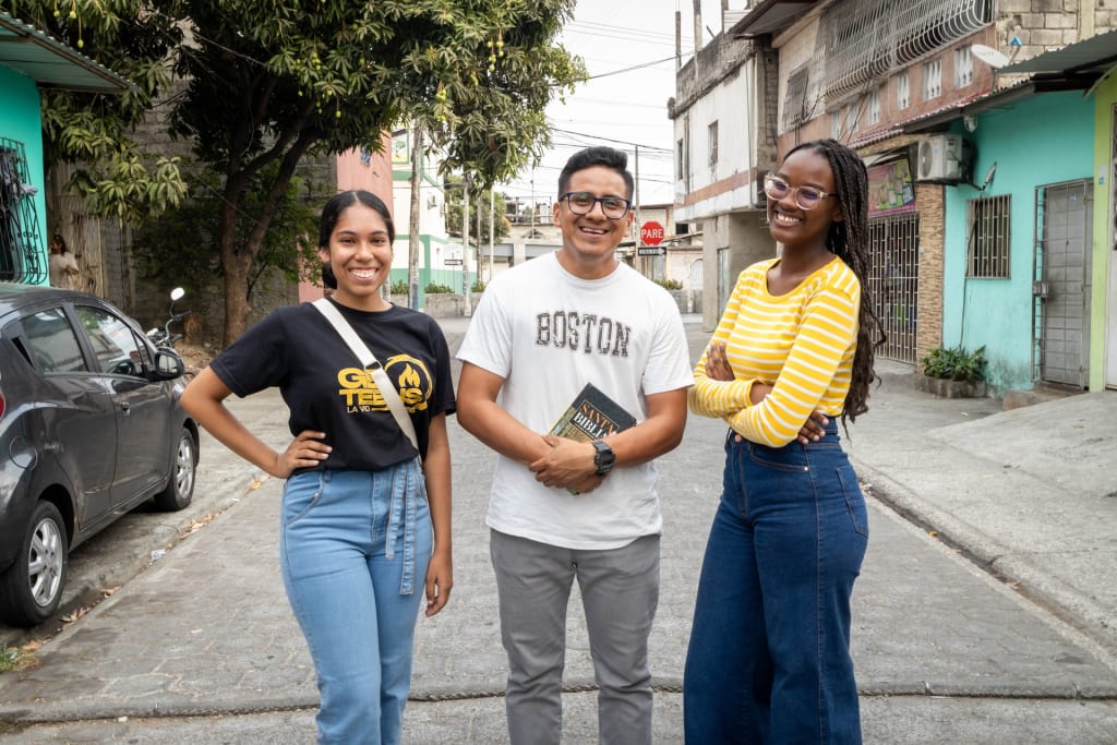 Tutor Sofia (in a black Tshirt and blue jeans), Pastor Daniel (in a white T-shirt that says BOSTON on it, and Fridy (in a yellow and white striped long-sleeved T-shirt and blue jeans) are standing together in the street smiling.