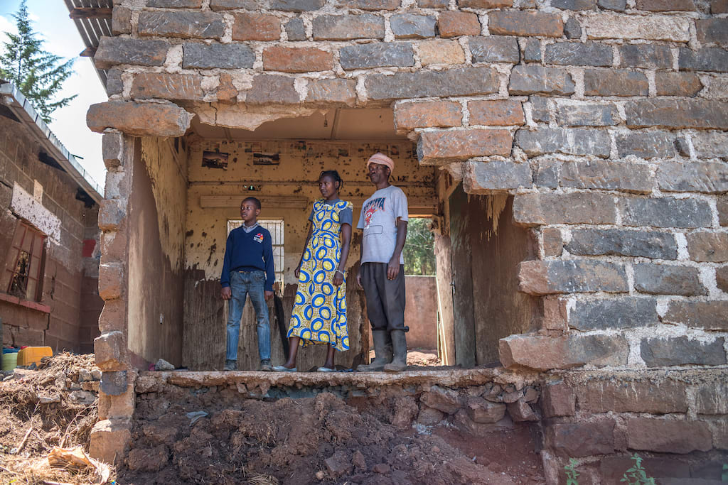 A family in Kenya stands in their destroyed home. Home Reconstruction is one of the gifts in the Gifts of Compassion gift guide.