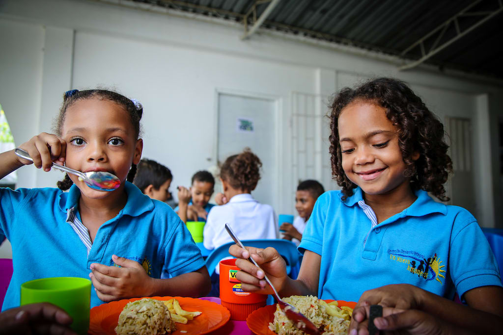 Two girls wearing blue shirts smile while eating food in a Compassion dining room.