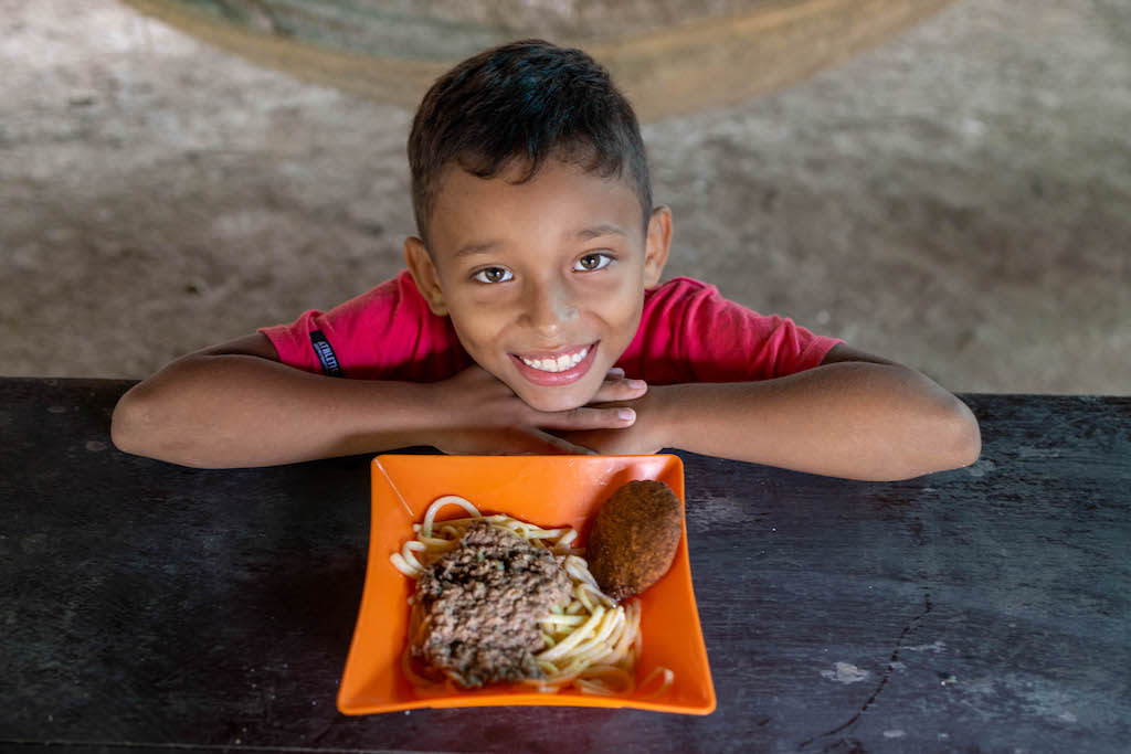 Little boy wearing a red shirt and smiles upward with a plate of food in front of him.