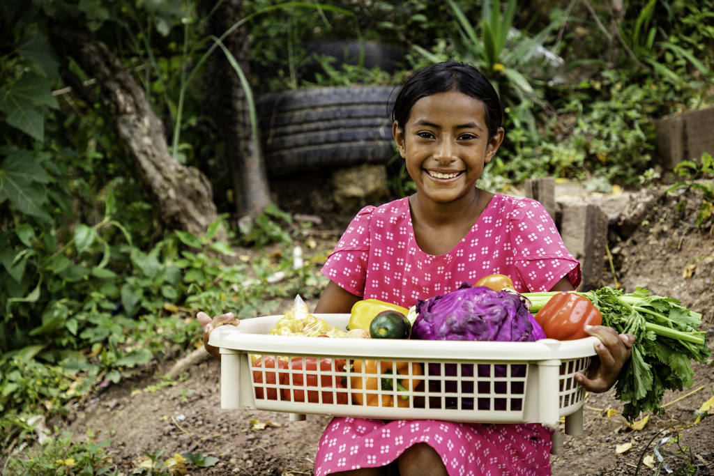 A girl in a pink dress with white polka dots holds a basket of vegetables.