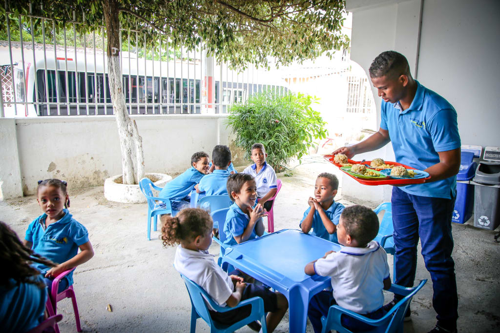 A man in blue serves a tray of food to children in blue sitting around small tables.