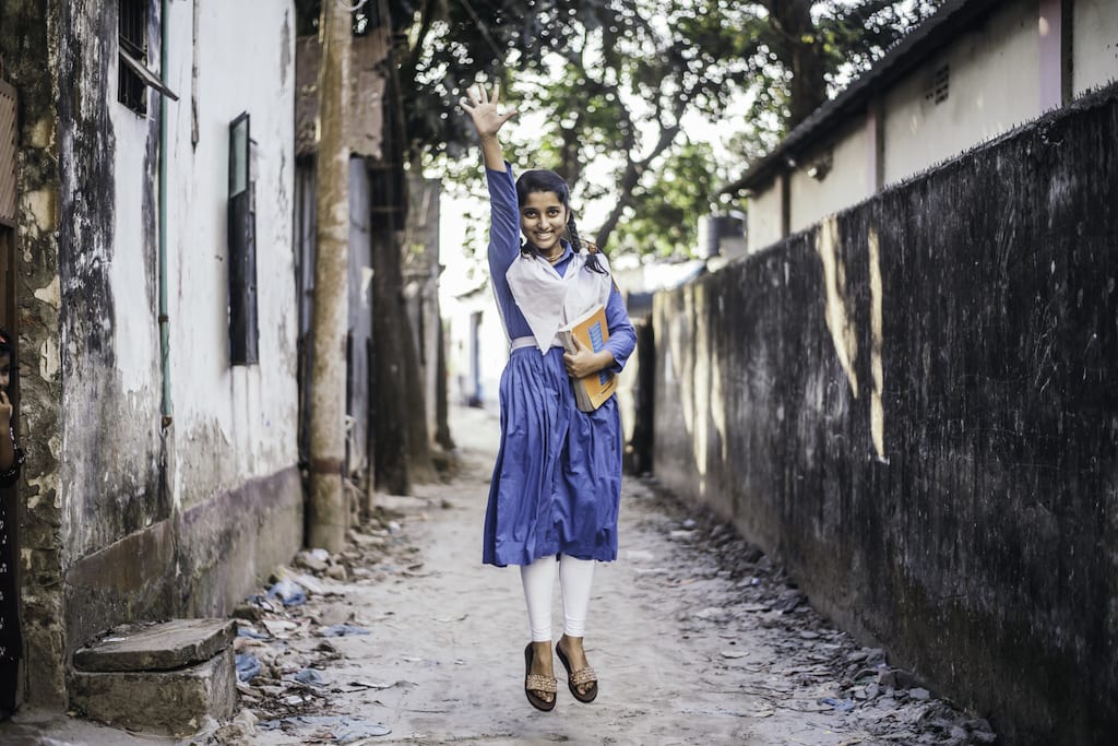 Tisha is wearing her school uniform, a blue dress with a white scarf. She is holding books and is jumping up ine air. She is standing in an alley.