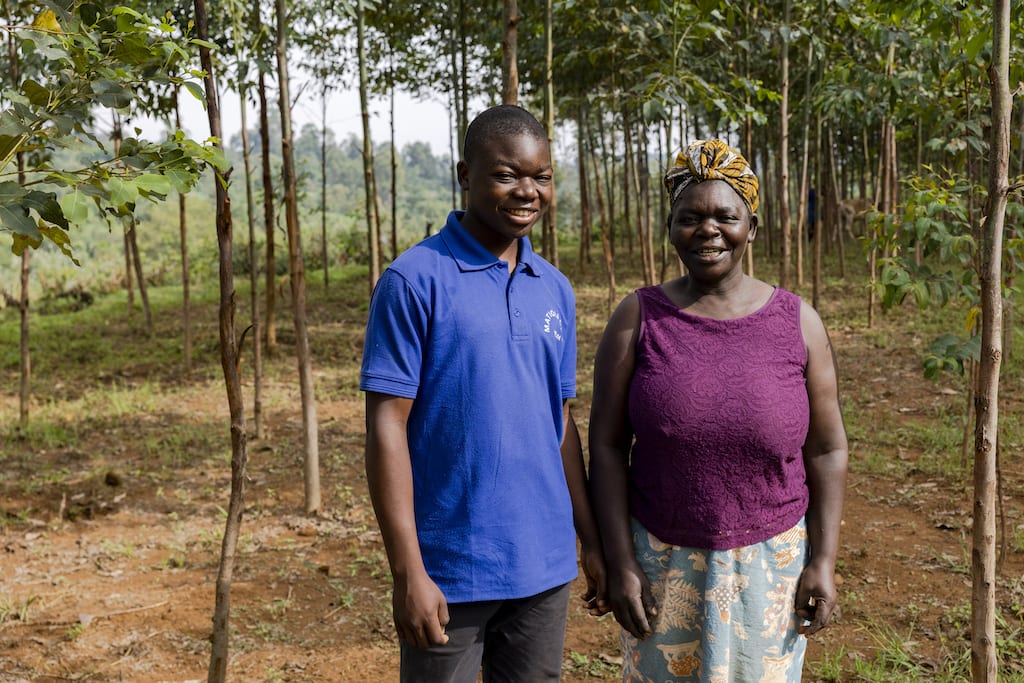 Jimson, wearing a blue shirt and black pants, is standing among trees he planted with his mother, wearing a maroon shirt and blue patterned skirt.