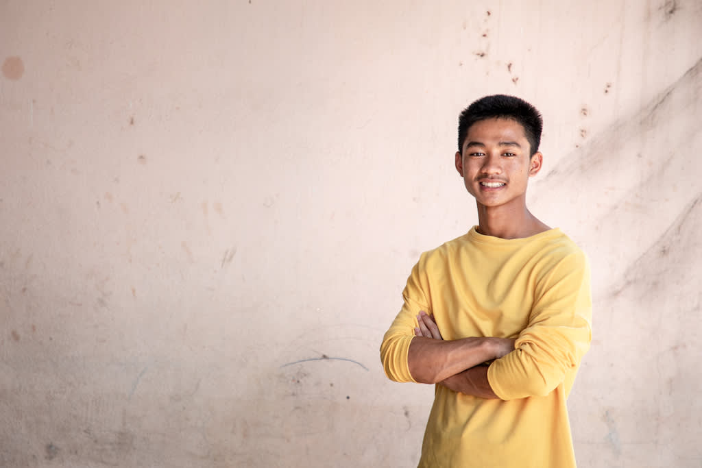 Adun, a member of the 'Thirteen Lives' soccer team, stands with his arms crossed. He is wearing a yellow long-sleeved t-shirt.
