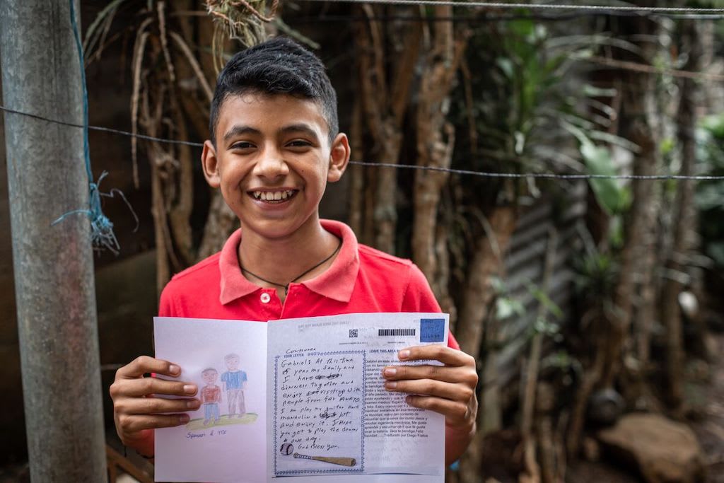In El Salvador, Gabriel is wearing a red shirt. He is standing in his back yard and is holding up a picture he drew for his sponsor and a letter from his sponsor. There are trees behind him.