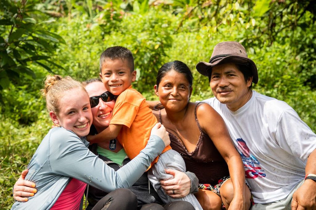 In Ecuador, Mallory, her mother Erin, Elian, and his family are smiling after Elian and his family got to meet their Compassion sponsor. There are trees in the background.