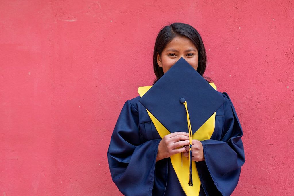 In Bolivia, Maribel is wearing her graduation uniform, a black gown and a yellow collar. She is standing in front of her red home and is holding her cap in front of her face.