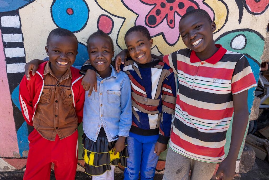 From left to right: John, Wamuyu, Dorcas, and Alphin, are standing together in front of a brightly colored mural. They have their arms around one another.