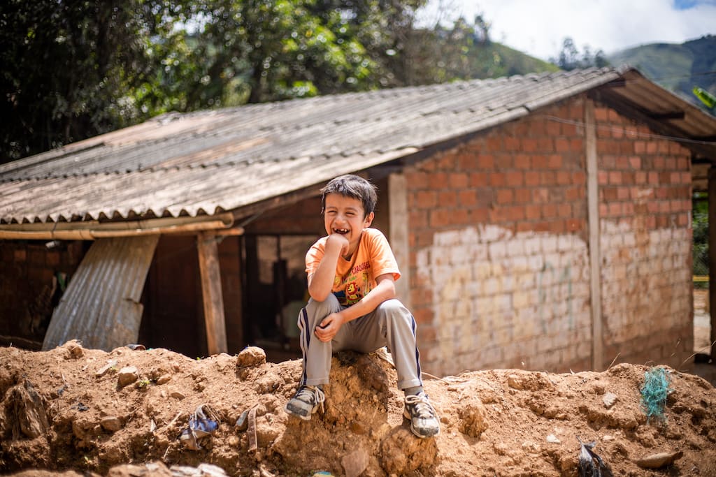A boy in Ecuador sits on the ground in front of his home. He is smiling and wearing an orange shirt.