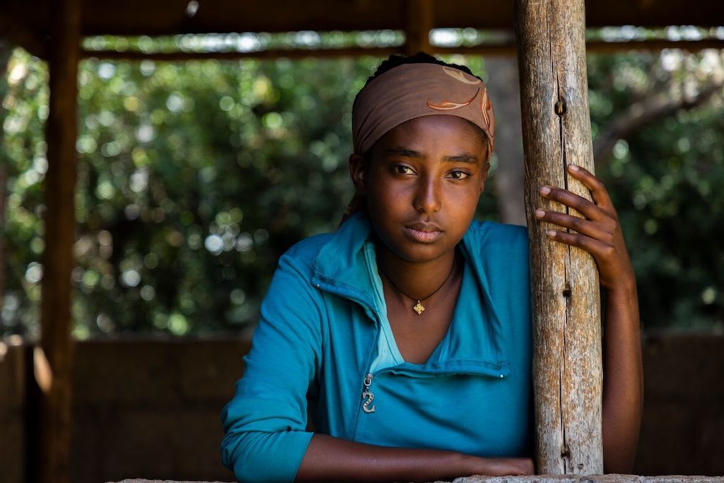 Bethlehem, a Ethiopian teenage girl, is wearing a turquoise blue shirt and a brown scarf around her head. She is sitting outside under a wooden shelter.