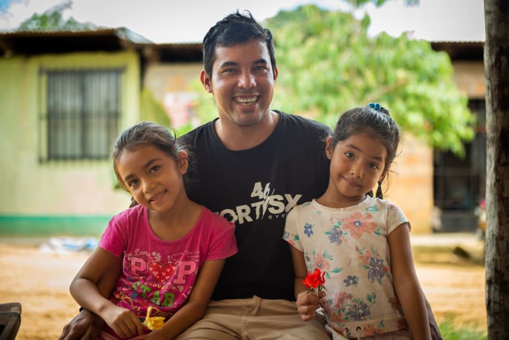 A father in a black shirt sits between his two girls wearing a pink shirt and a floral top. They are smiling.