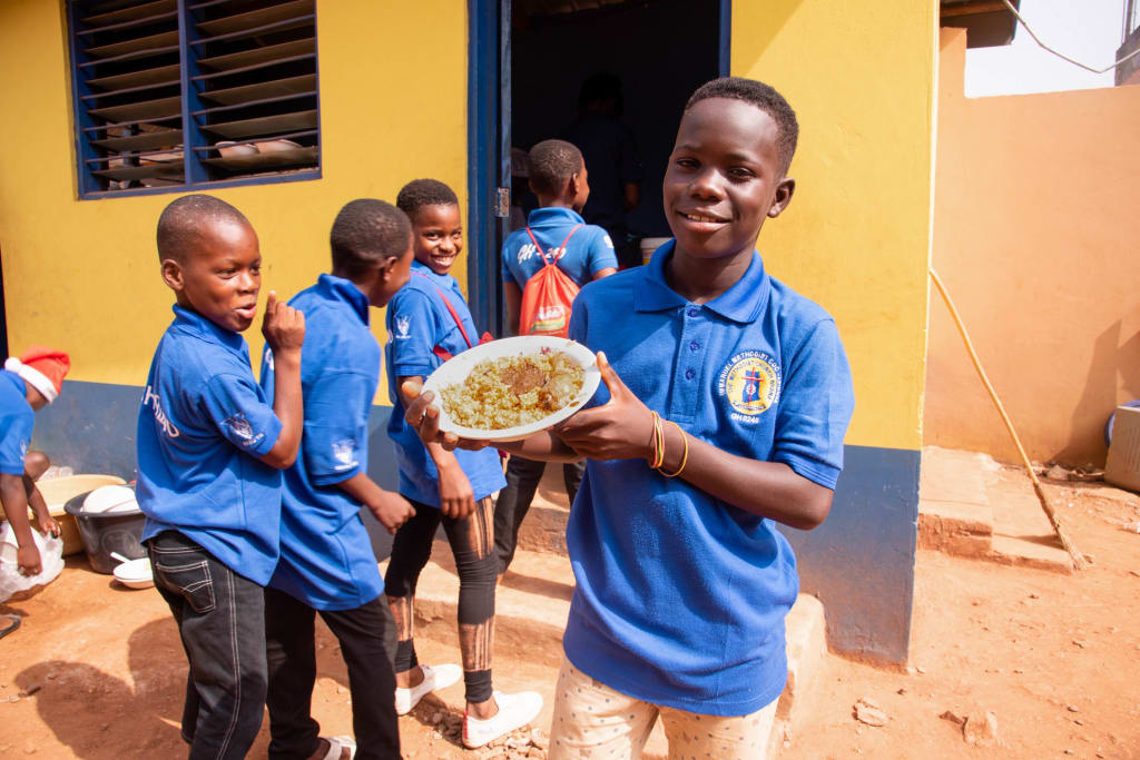 Young African boys stand around in school uniforms as one holds up a plate of food