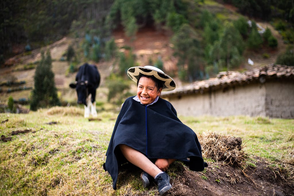 In Ecuador, 11-year-old Davis is wearing traditional clothing. He is sitting in a field outside his home. There is a cow behind him.