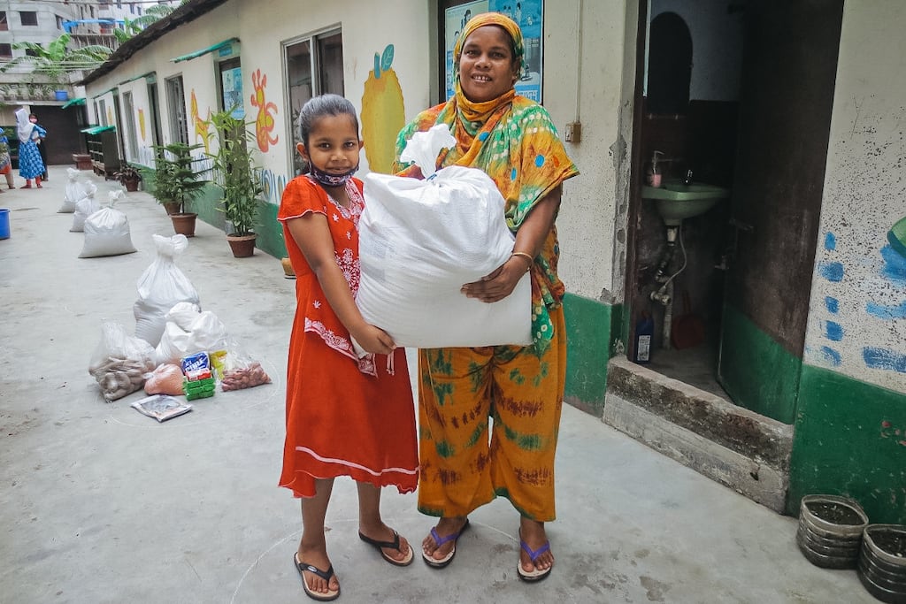 Mim is wearing an orange dress. She is standing with her mother, who is wearing a yellow, green, and brown outfit. Together, they are holding a white plastic bag full of food they received from Compassion. The Compassion building behind them is painted with a colorful mural.