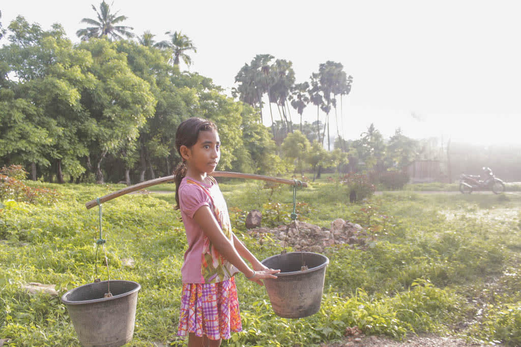 Yunita stands in a green field wearing a pink outfit and carrying a yoke with two water buckets balancing on her shoulder.