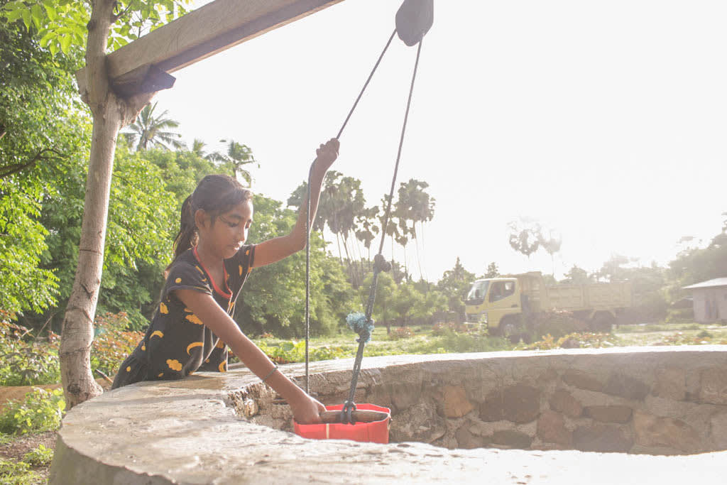 Wearing a black outfit, Yunita collects water from a large well using a red bucket.