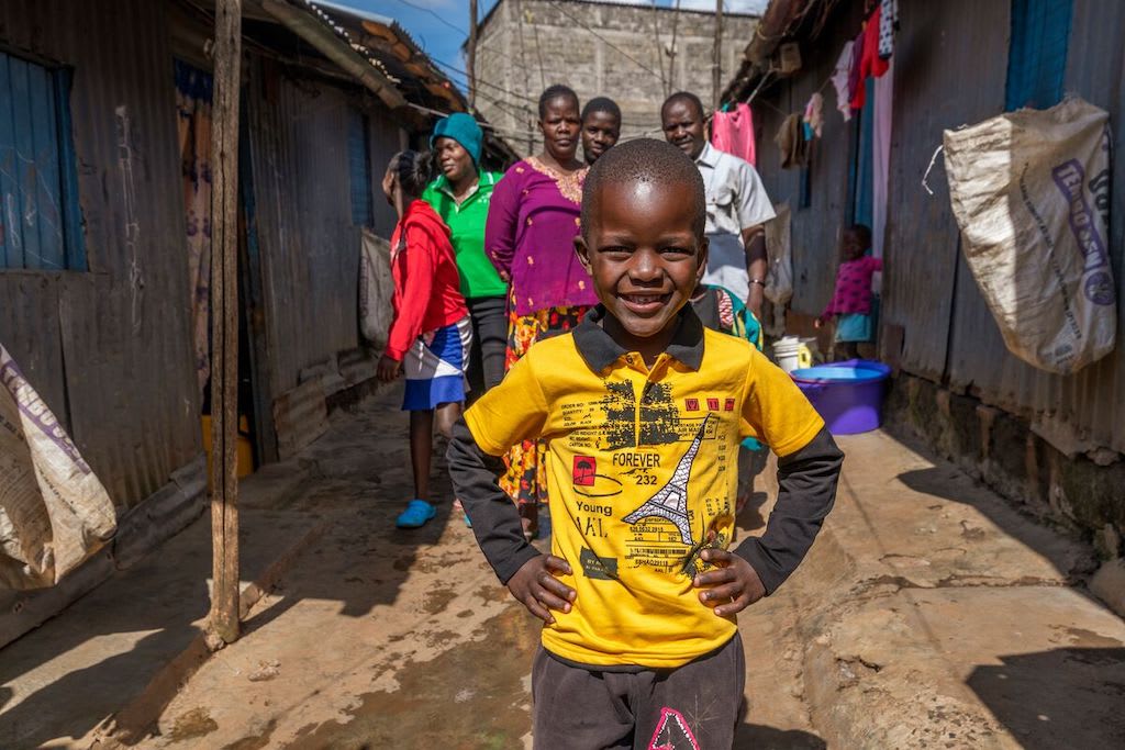 In Kenya, a boy in a yellow shirt stands with his hands on his hips, with his family standing behind him.