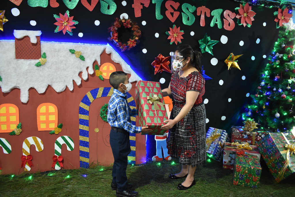 A woman wearing a mask passes a gift to a little boy wearing a mask. There is a painted gingerbread house in the background.