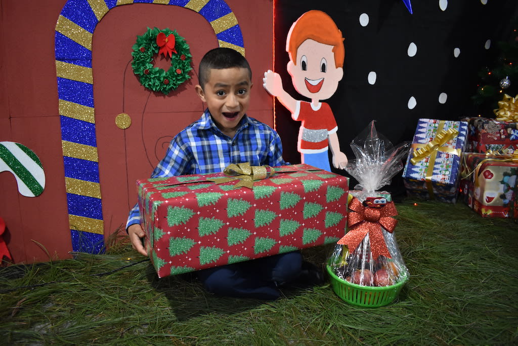 Little boy wearing a blue plaid shirt sits with a big gift on his lap, with a big happy surprised look on his face.