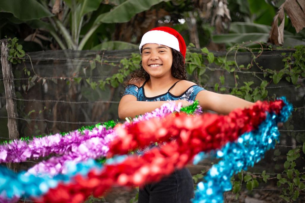 Young girl stands infront of trees wearing a santa hat and golding colourful tinsel.