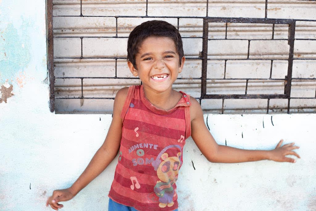 A child from Pastor Linaldo's community stands against a wall, smiling and wearing a red shirt.