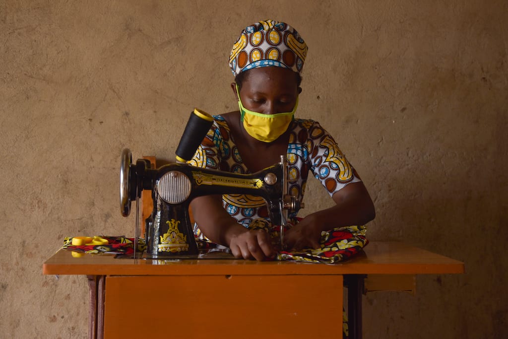 In Rwanda, Alice, the mother of two Compassion sponsored children, is wearing a colorfully patterned dress and a yellow face mask. She is sitting at a sewing machine in her shop and is sewing a dress.