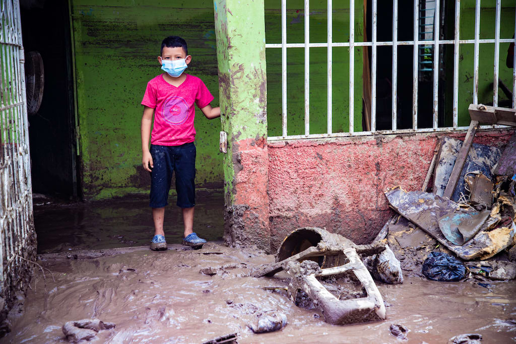 A boy in a face mask and a red shirt stands in the green doorway of his home. The floor and walls are muddy.