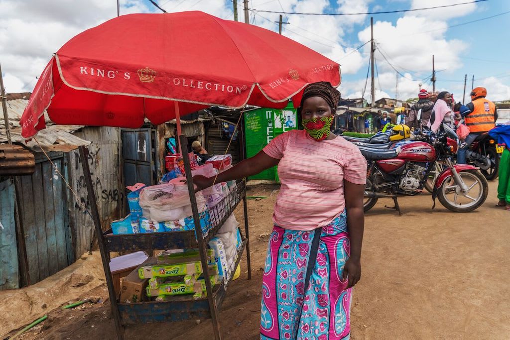 A woman in a pink shirt and patterned face mask stands at a stall under a red umbrella, selling consumer items.