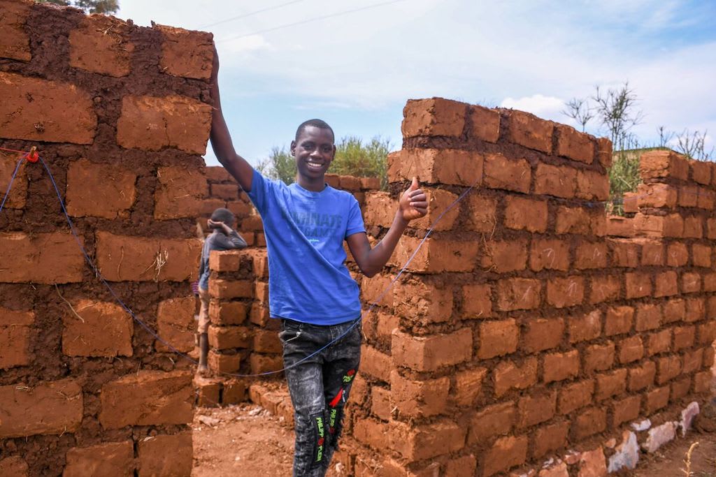 A teenage boy in Rwanda stands amongst unfinished brick walls that will make up his family's new home.