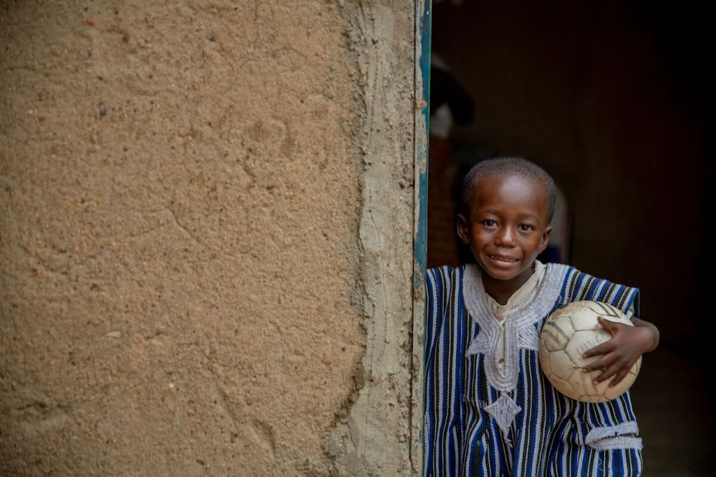 Achille stands in a doorway holding a soccer ball under his arm.
