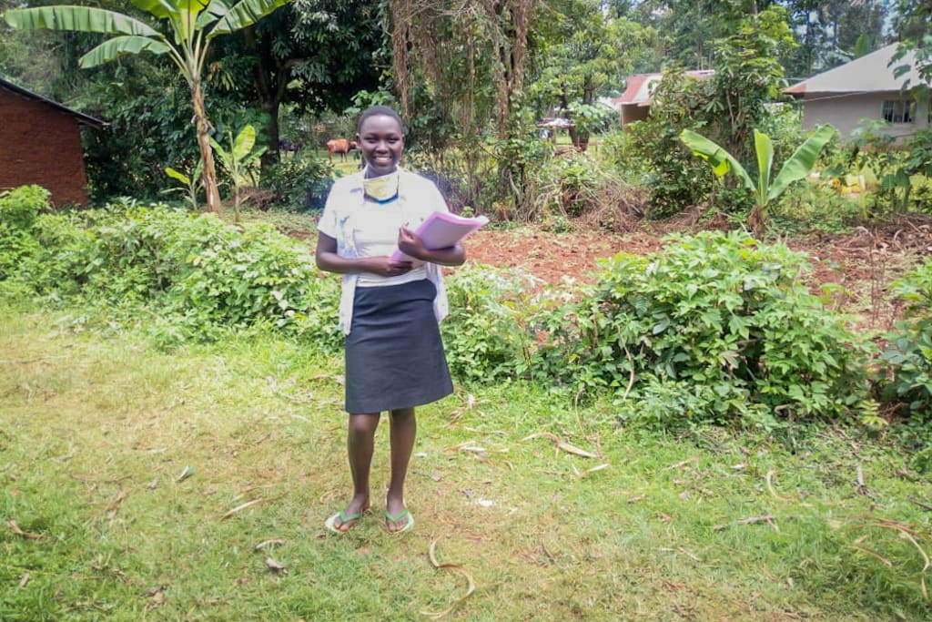Sharon stands in a field outside her Compassion centre, holding her school books. She has a protective facemark on her chin.