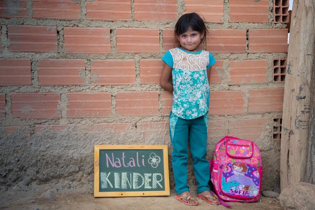 Natali stands against a brick wall with her pink backpack next to her and a chalkboard that says "Natali KINDER" on her other side.