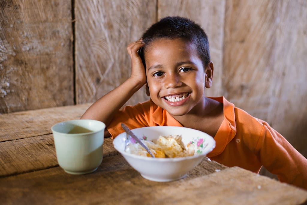A boy smiles with a bowl of food on the table in front of him.