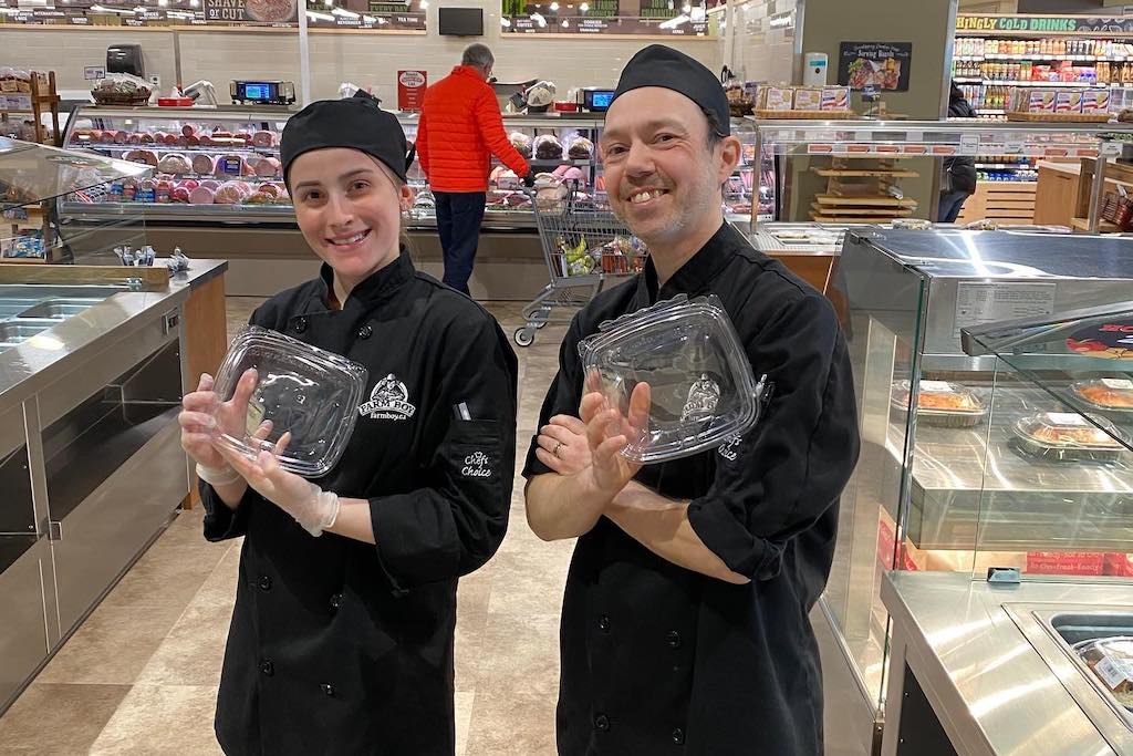 Two grocery store employees stand with takeout containers.