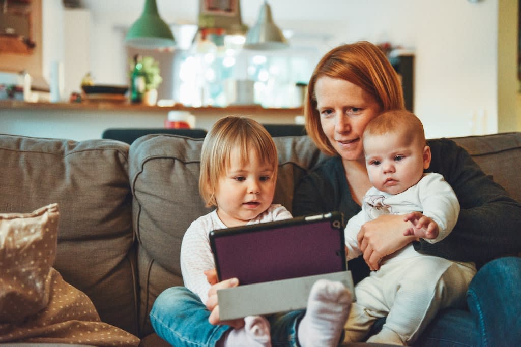 A woman looking at a tablet with a baby and a toddler, sitting on a couch.